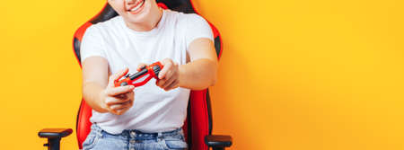 Woman sitting behind gaming chair in her hands holding red gamepad on yellow background.