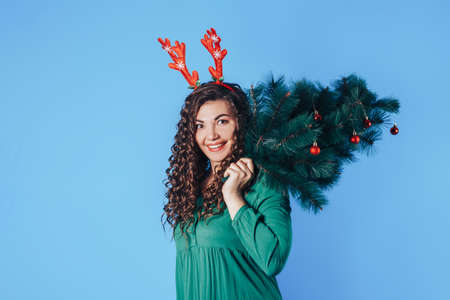 Young beautiful woman on yellow background, she has antlers on her head. Woman holding Christmas tree on her shoulder. Christmas mood. Place for text.の写真素材