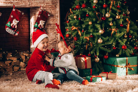 Brother and sister are sitting with boxes in green packaging with gifts and a bow. Christmas mood. Children in New Year's costumes waiting for a miracle. Against the background of a Christmas tree and fireplace.の写真素材
