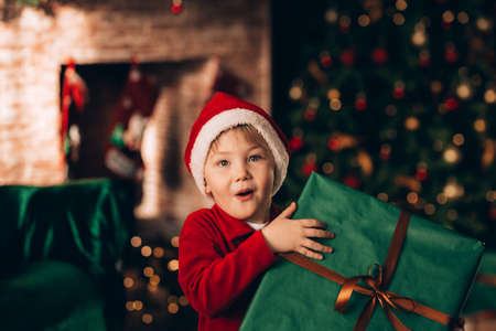 A boy holding a large box in a green package and a bow with a gift. Christmas mood. Against the background of a large Christmas tree and a fireplace.の写真素材