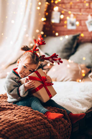 A little girl holding a large gift in a paper box on a large bed. The child has antlers on his head. Christmas mood.の写真素材