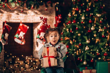 Little girl sitting in front of a decorated Christmas tree. Against the background of a Christmas tree with boxes in green packaging with a bow and a fireplace. Christmas moodの写真素材