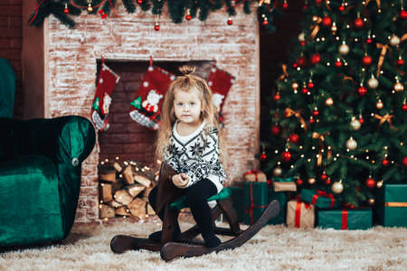 Child sits on a wooden swing in the shape of a horse. Christmas mood. In the background a decorated Christmas tree and a large fireplace.の写真素材