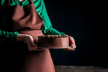 Woman baker in apron holding a chocolate cake on a dark background. Workers uniform in the kitchen. Place for text.の写真素材
