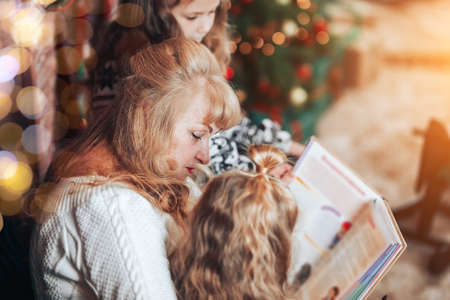 Grandmother with children read holding a book in their hands. Christmas mood. In the background there is a fireplace and a green armchair.の写真素材