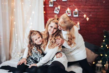Child with mom and grandmother are seated hugging on the big bed. Christmas mood. Against the background of a brick wall with garlands.の写真素材