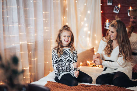 Mom and daughter are sitting on the big bed. Christmas mood. Against the background of a brick wall with garlands.の写真素材