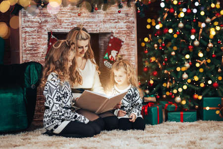 Mom with children read holding a book in their hands. Christmas mood. In the background there is a fireplace and a green armchair and a Christmas tree.の写真素材