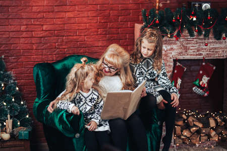 Grandmother with children read holding a book in their hands. Christmas mood. In the background there is a fireplace and a green armchair.の写真素材
