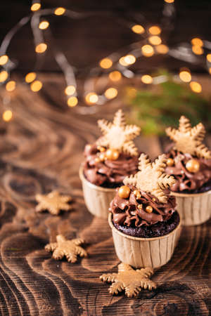 Chocolate cupcakes with star shaped cookies on a wooden background. Christmas mood. Sweets for any occasion. In the background are yellow lights from the garlandの写真素材