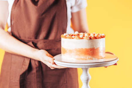 Woman baker holding cake on a stand. Uniforms of kitchen workers in front of a yellow background....