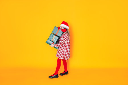 Child holding large boxes with gifts. There is a Santa hat on her head, and a medical mask on her face. Christmas mood. A perfect photo for a Christmas advertisement during a pandemic.の写真素材