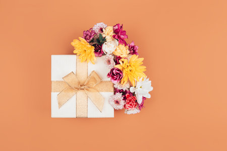A mix of various flowers next to a beautiful white box wrapped with a ribbon, a beautiful bow on the lid of the box, a gift for March 8 on an orange backgroundの写真素材