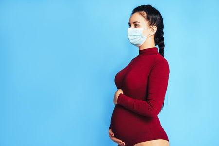 Pregnant woman in front of a blue background. The woman is wearing a medical protective mask. Caring for the health of a pregnant woman.の写真素材