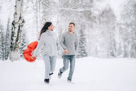 A loving couple run through the snow in the forest. Woman holding heart-shaped balloons in her hands.の写真素材