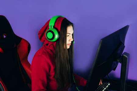 A young girl playing a computer game on professional armchair against monitor in green headphones,...