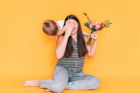 A little boy gives his mother a bouquet of flowers and covers her eyes with his hand against a yellow backgroundの写真素材