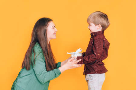 Little boy gives mom a box with a gift against a yellow background. Gift for womens day and mothers dayの写真素材