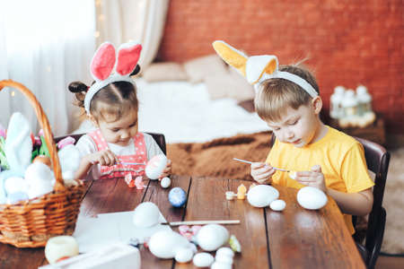 A boy and a girl are painting Easter eggs for the holiday at a wooden table. Children have bunny ears on their headsの写真素材