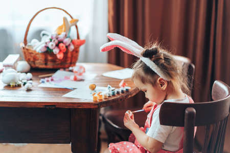 Girl are painting Easter eggs for the holiday at a wooden table. Child have bunny ears on their...