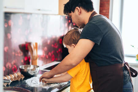 Father and son roll out the dough with rolling pin. Preparing for the Mothers Day holiday. Asian father and mestizo son prepare food in the kitchenの写真素材