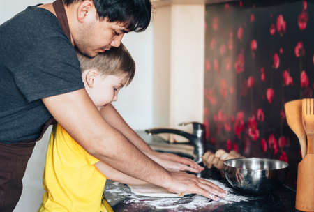 Father and son roll out the dough with rolling pin. Preparing for the Mothers Day holiday. Asian father and mestizo son prepare food in the kitchenの写真素材