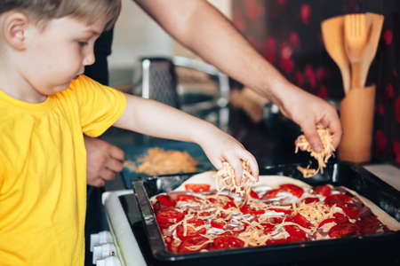 Father and son sprinkle grated cheese on pizza. Preparing for the Mothers Day holiday. Father and son preparing food in the kitchenの写真素材