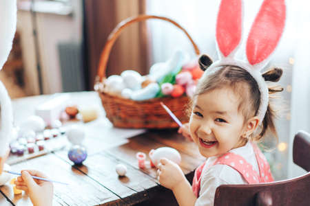 Preparing for Easter, child paints eggs on wooden table for religious holiday. Girl dressed in bunny...