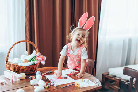 Preparing for Easter, child paints eggs on wooden table for religious holiday. Girl dressed in bunny costume.の写真素材