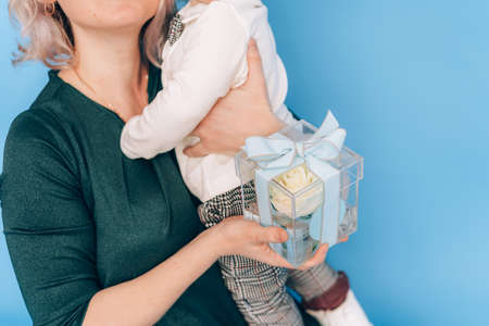 Woman in dress holding little boy in her arms and holding beautiful flower in against blue background. Holiday gift.の写真素材