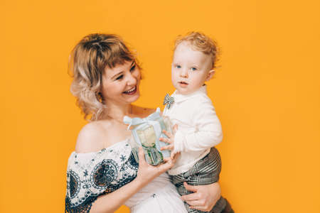Woman in dress holding little boy in her arms and holding beautiful flower in against yellow background. Holiday gift.の写真素材