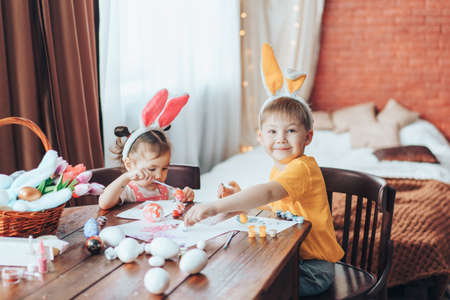 Preparing for Easter, children paint eggs for religious holiday. Boy and girl are painting Easter eggs for the holiday at wooden table. Children have bunny ears on their heads.の写真素材