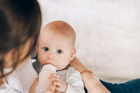 Young mother is holding her newborn baby in her arms and feeding from bottle. Love and care for children. Adoption of children. The child grows up without a fatherの写真素材
