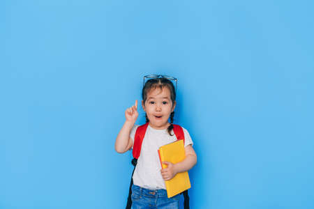Surprised schoolgirl holding yellow book in front of blue background. Back to school in 2021. Homeschooling, distance learningの写真素材