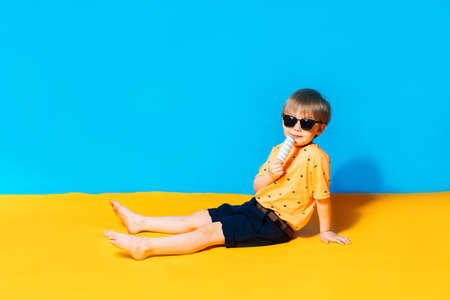 Summer and vacation in 2021. Mixed race boy in sunglasses licks an ice cream. The blue background...