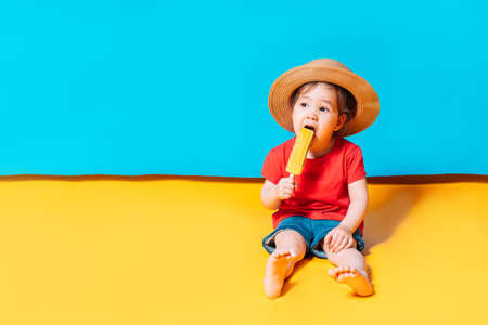 Asian girl in a straw hat licks ice cream. The blue background shows the sky, and the yellow background shows the sand.の写真素材