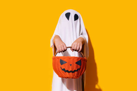 Child in ghost costume holding pumpkin-shaped basket in front of him.の写真素材