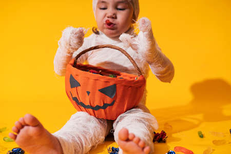 Child in mummy costume eats candies from basket in the form of pumpkin against yellow background. Halloween trick or treatの写真素材