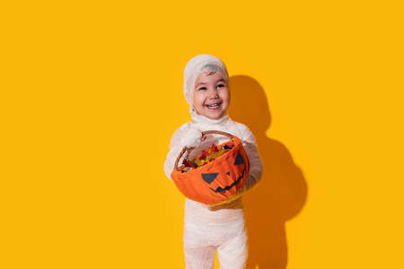 Child in mummy costume holding basket of chocolates in front of yellow background.の写真素材