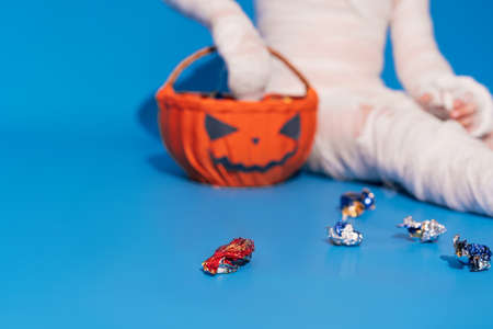 Child in mummy costume eats candies from basket in the form of pumpkin against blue background. Halloween trick or treatの写真素材