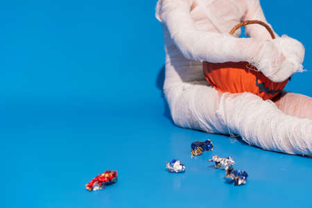 Child in mummy costume eats candies from basket in the form of pumpkin against blue background.の写真素材