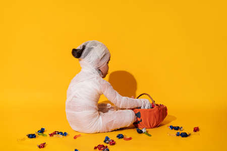 Child in mummy costume eats candies from basket in the form of pumpkin against yellow background.の写真素材