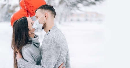 Young couple in love in the park on a background of snow and red heart-shaped balloons. Valentine's day gift. Lovers kiss in medical masks.の写真素材