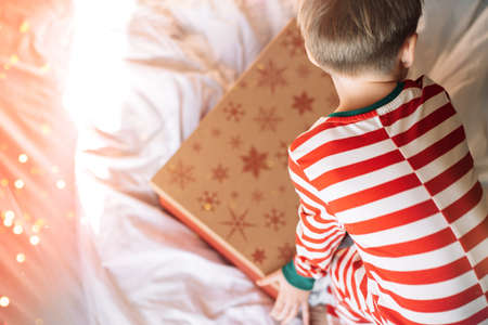 Boy in striped pajamas closes gift box on white bed with superimposed lightsの写真素材