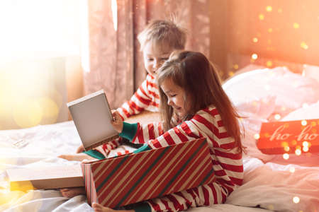 Children in striped pajamas unpack Christmas gifts while sitting on white bed.の写真素材