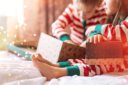 Children in striped pajamas unpack Christmas gifts while sitting on white bed.の写真素材