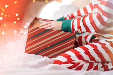 Child in striped pajamas unpacks gift box on white bed with superimposed lights.の写真素材
