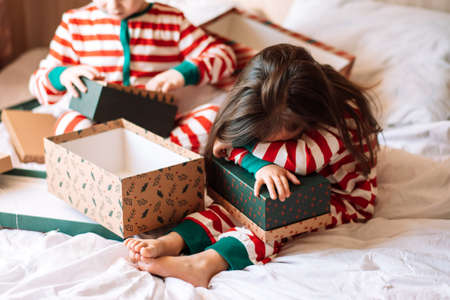 Siblings in pajamas unpack Christmas gifts while sitting on white bed.の写真素材