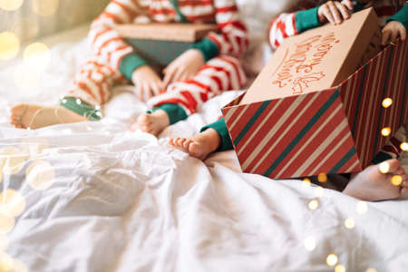 Children in striped pajamas unpack Christmas gifts while sitting on white bed.の写真素材