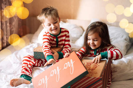Children in striped pajamas unpack Christmas gifts while sitting on white bed.の写真素材